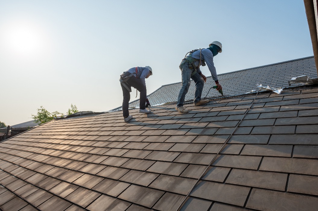 Ein Dachdecker, der spezielle Schutzhandschuhe trägt und mit einem luftbetriebenen oder pneumatischen Nagler arbeitet, repariert und ersetzt Dachziegel auf dem neuen Dach eines im Bau befindlichen Wohnhauses